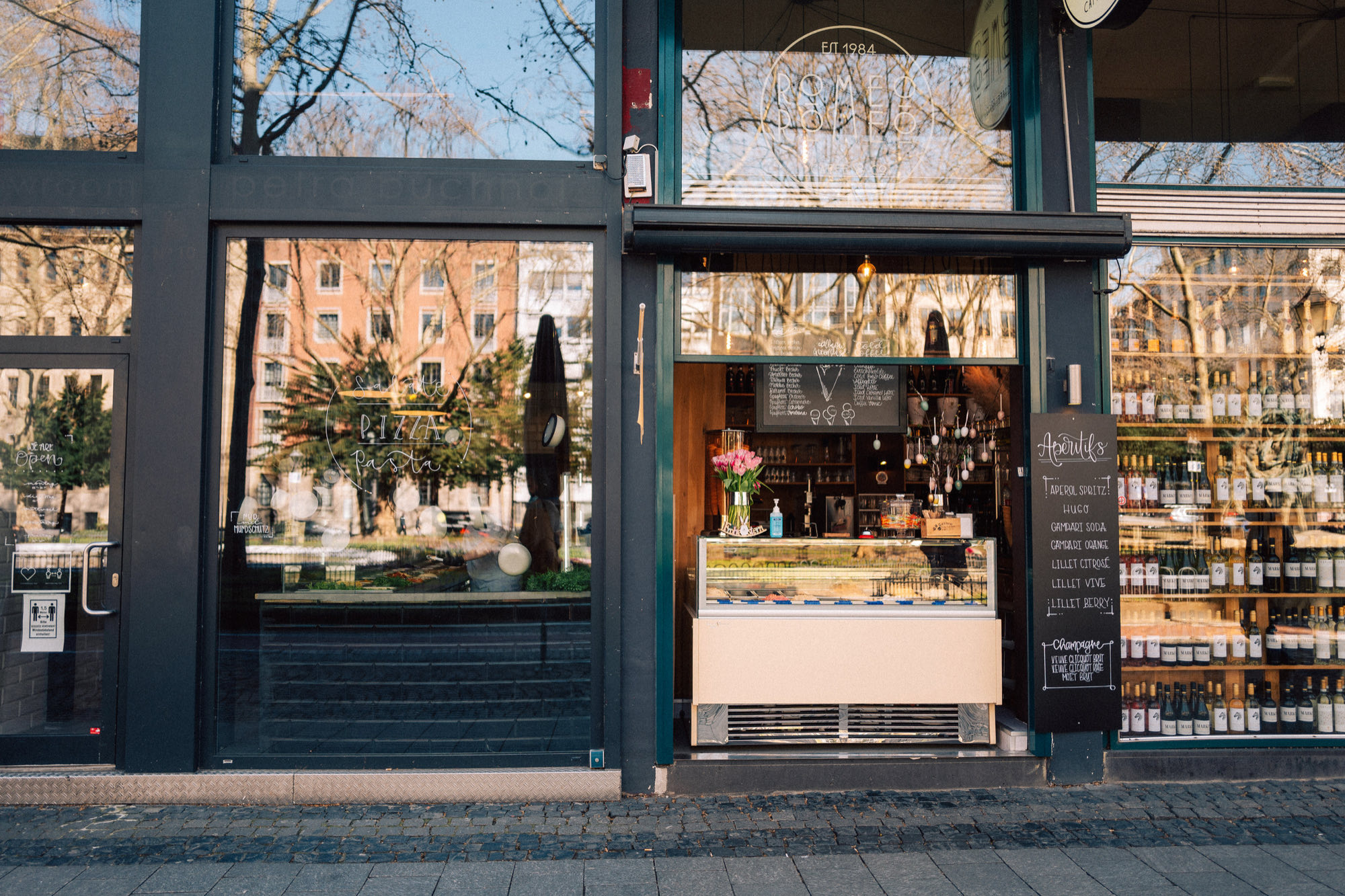 Schaufensterfront des Cafés von der Straße aus, dahinter die Kuchenvitrine und eine beschriebene Tafel